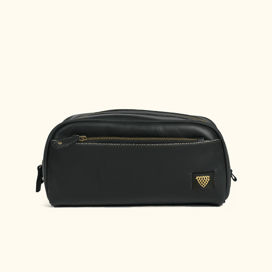 A black leather cosmetic pouch with gold-tone hardware and a small triangular gold logo patch on the front, set against a solid cream background.