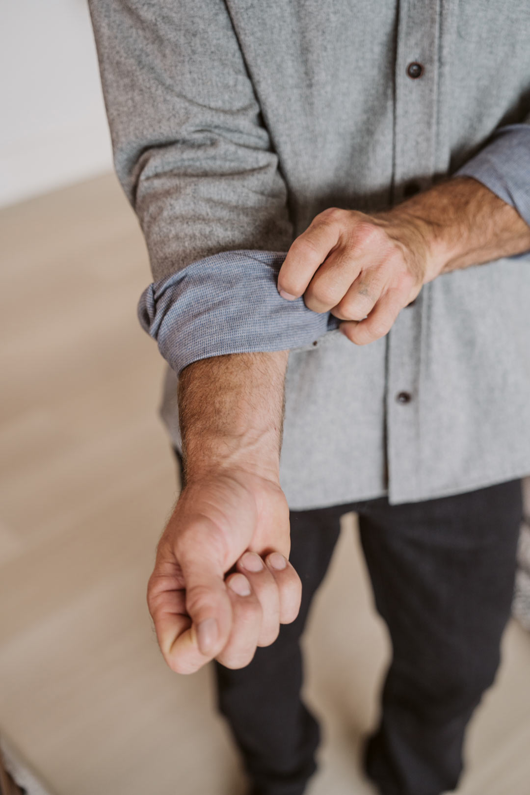 A close-up of a man's hands rolling up the sleeve of a heather gray long-sleeve button-up shirt, paired with dark trousers, in a brightly lit indoor setting.