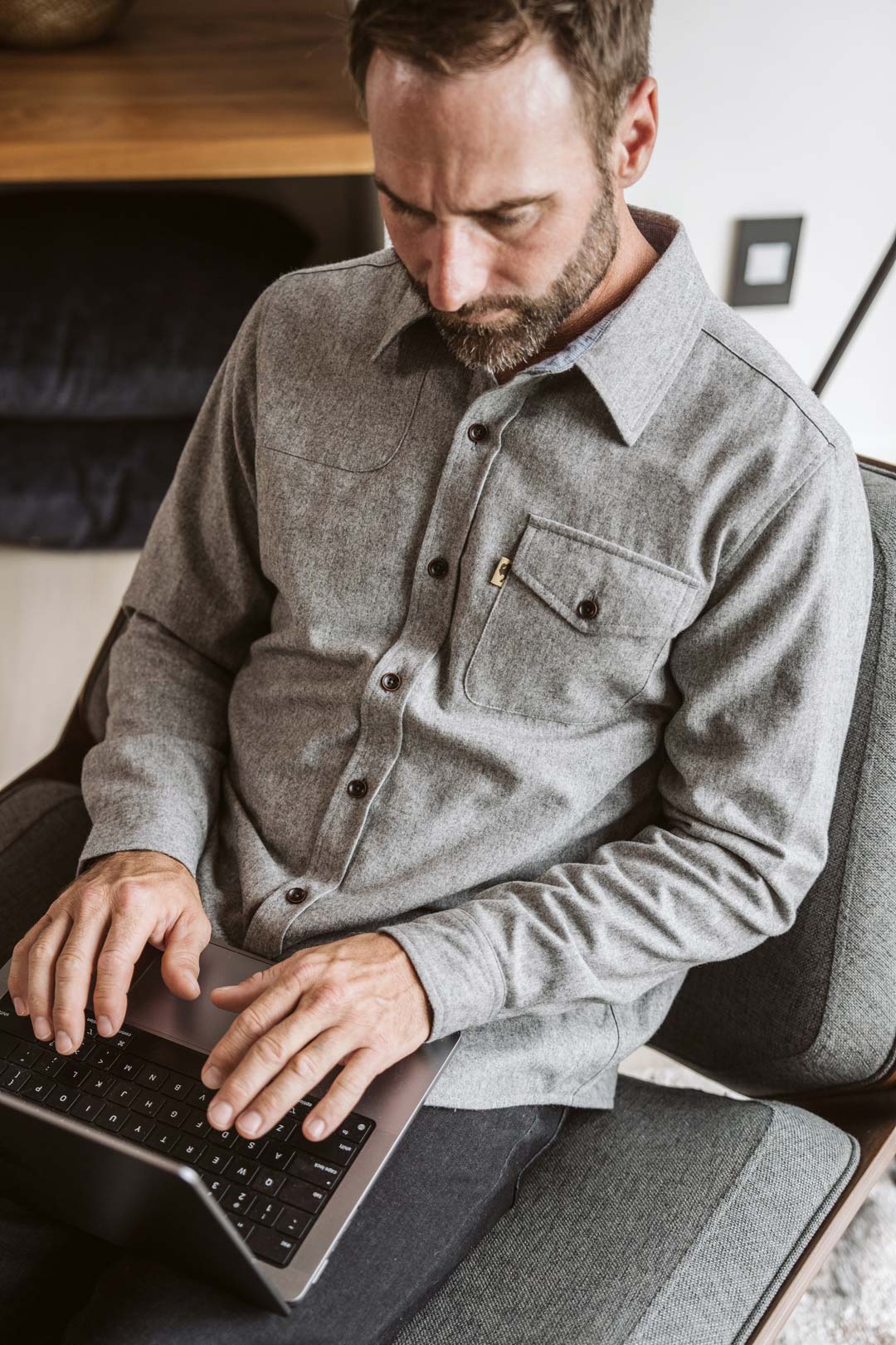 A man wearing a gray long-sleeve button-up shirt sits in a chair and types on a laptop in a modern indoor setting.