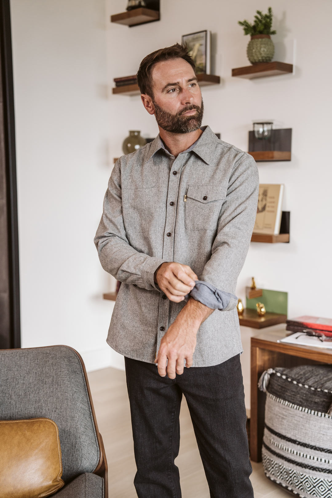 A man wearing a light gray long-sleeve button-down shirt and dark pants, standing in a modern living room setting.