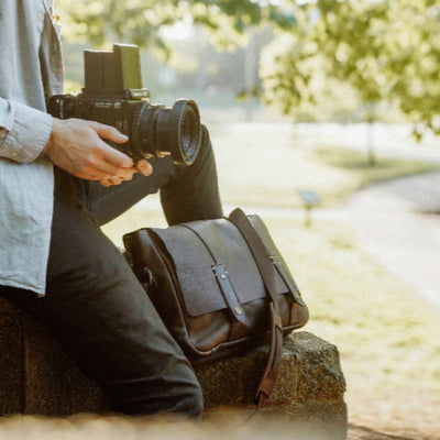 Person sitting outdoors with a camera and bag, surrounded by greenery