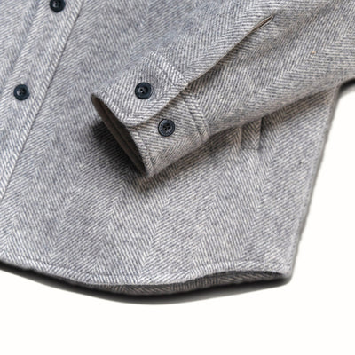 Close-up of a gray herringbone patterned shirt jacket or overshirt with dark buttons, showing the fabric texture and cuff detail against a white background.