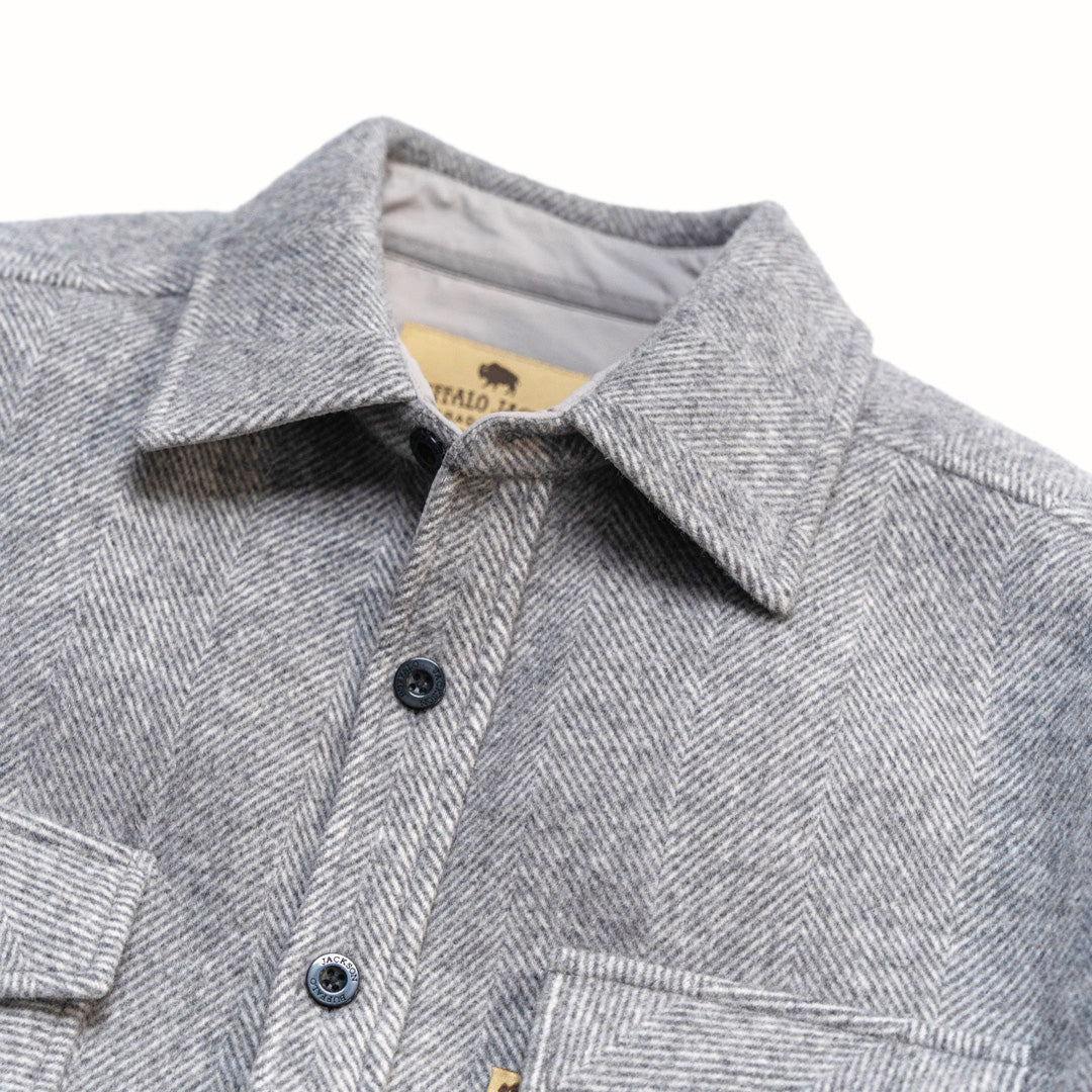 Close-up of a gray herringbone pattern button-down shirt jacket with a pointed collar and dark buttons, showing a tan neck label with buffalo branding.