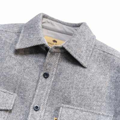 Close-up of a gray herringbone pattern button-down shirt jacket with a pointed collar and dark buttons, showing a tan neck label with buffalo branding.