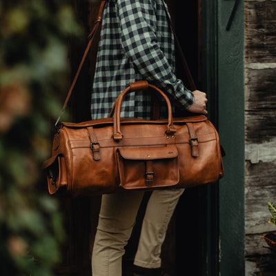 man is holding a Roosevelt Buffalo Leather Duffle Bag in Autumn Brown. He is casually dressed in a plaid shirt and light khaki pants, standing in a doorway, ready to step out or travel, with the bag slung over his shoulder.