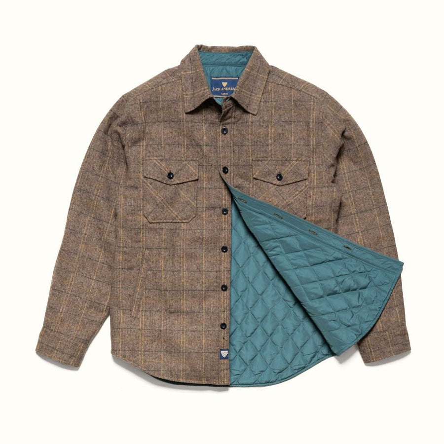 A man wearing a brown textured tweed button-up shirt and blue jeans, sitting on a wooden stool against a neutral beige fabric backdrop.