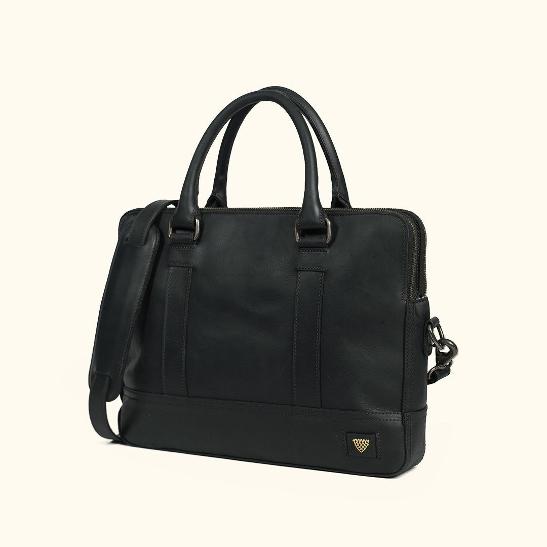 A black leather briefcase with dual top handles, a detachable shoulder strap, and antique nickel hardware, with a small gold shield logo on the bottom right corner, set against a solid cream background.