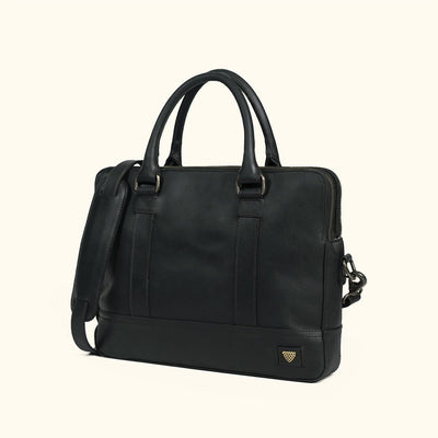 A black leather briefcase with dual top handles, a detachable shoulder strap, and antique nickel hardware, with a small gold shield logo on the bottom right corner, set against a solid cream background.