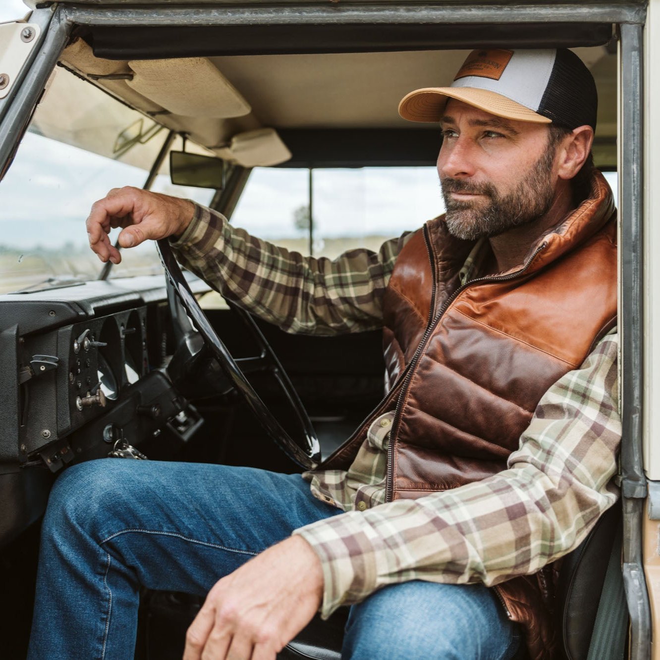 Man seated in a vintage truck wearing the Bridger Leather Down Vest in Tan and Brown