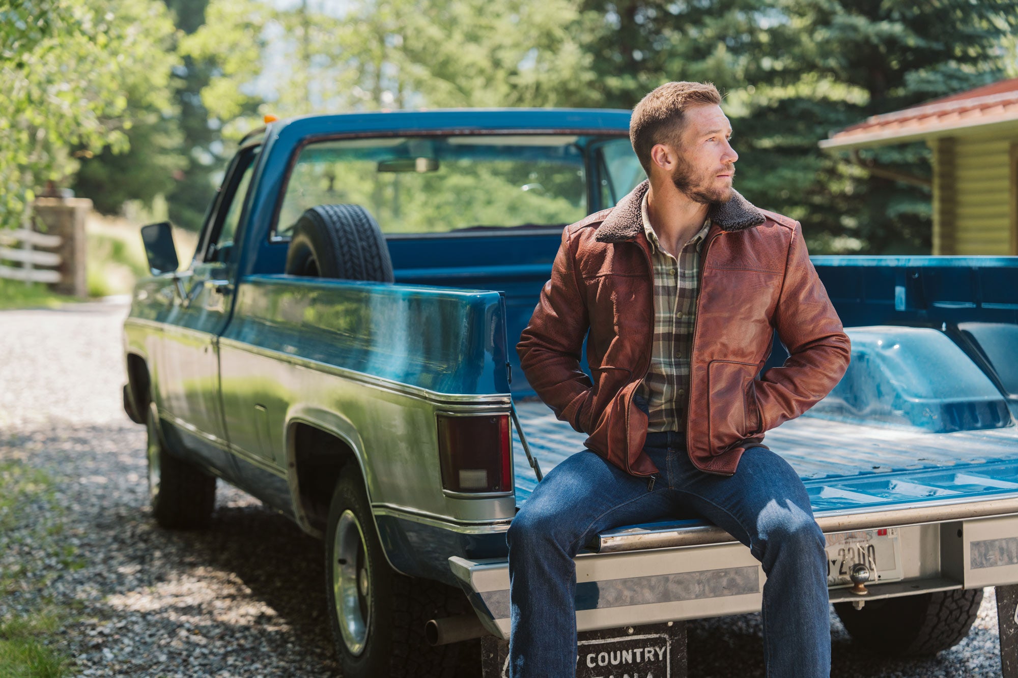 Man sitting on a pickup tailgate wearing the Limited Maverick Leather Bomber Jacket in Buffalo Grain Brown over a plaid flannel