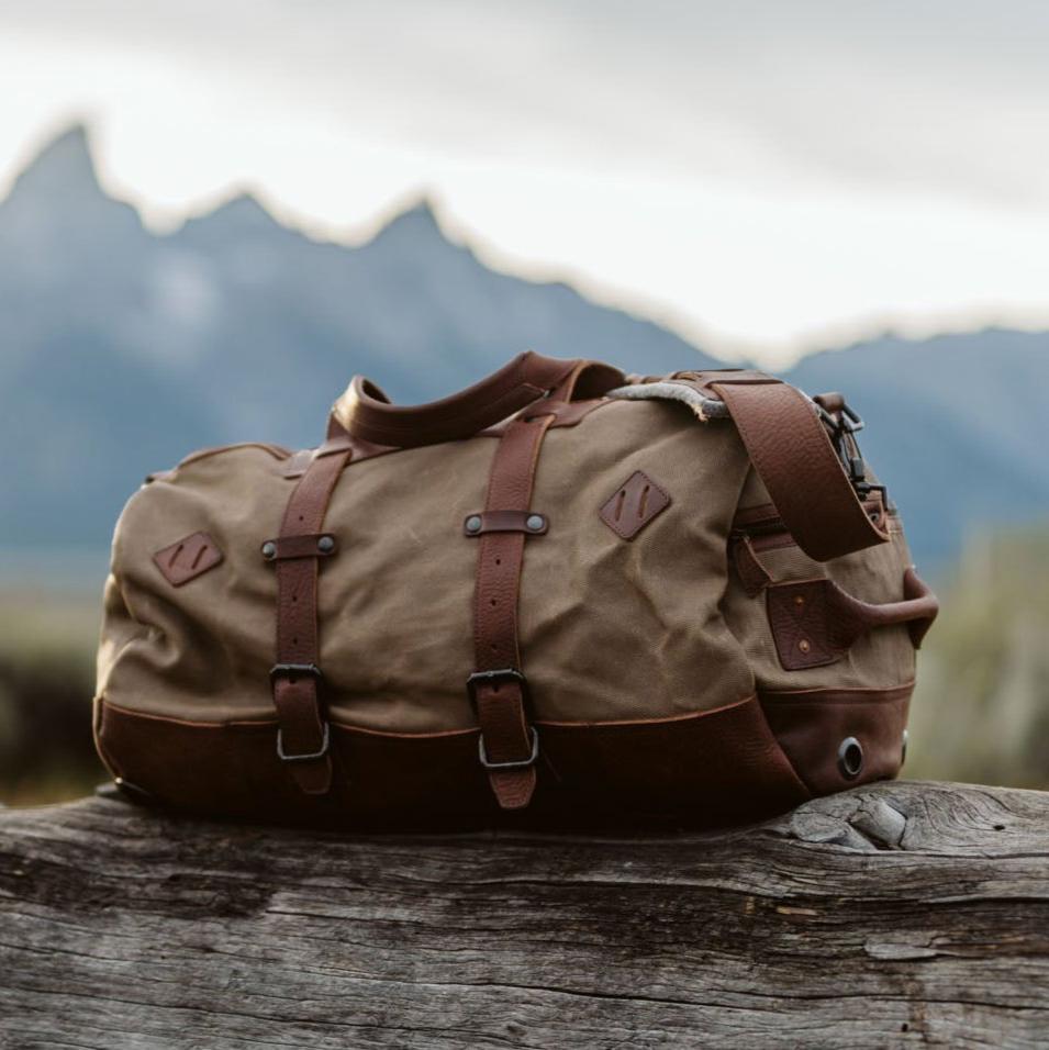 A waxed canvas duffle bag with chestnut brown leather accents, conversion straps, and a field khaki color, displayed on a wooden surface with a mountainous backdrop.