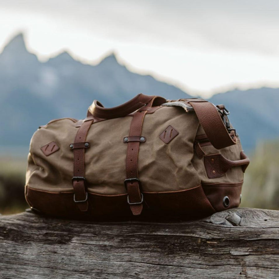 A waxed canvas duffle bag with chestnut brown leather accents, conversion straps, and a field khaki color, displayed on a wooden surface with a mountainous backdrop.
