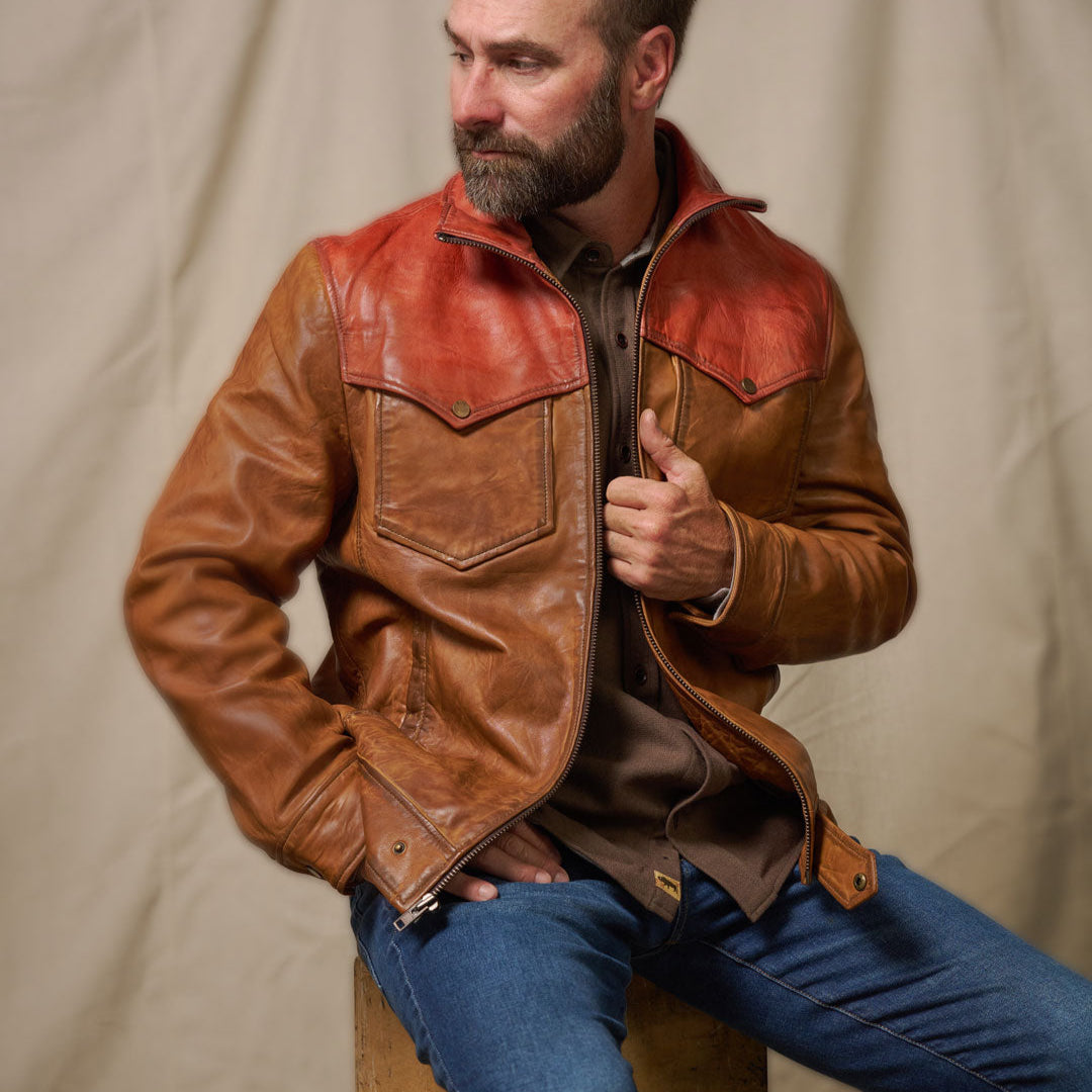 Man wearing a brown western leather jacket and blue jeans sitting on a wooden stool against a beige background