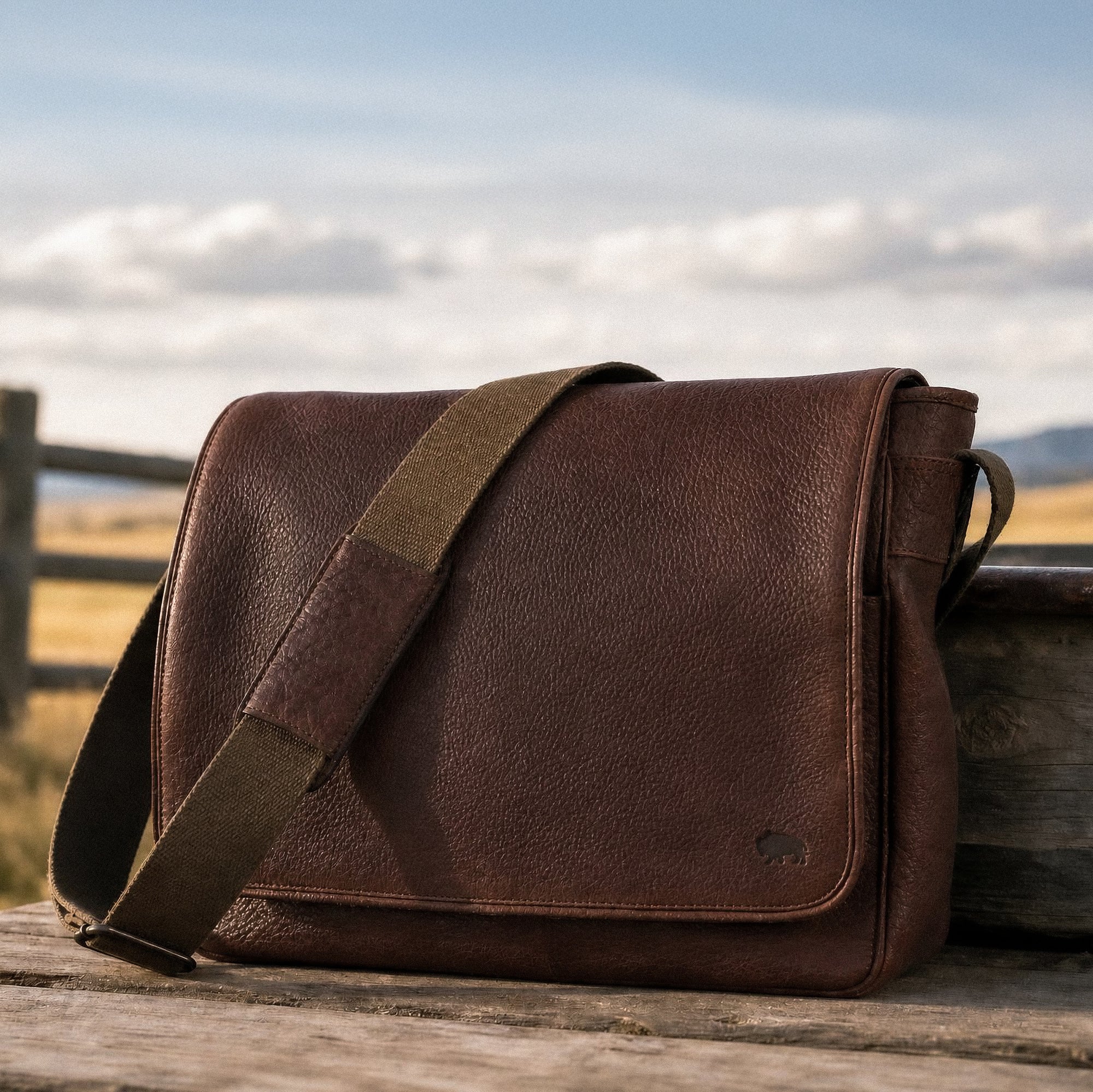 Brown leather messenger bag on a wooden surface with a scenic background