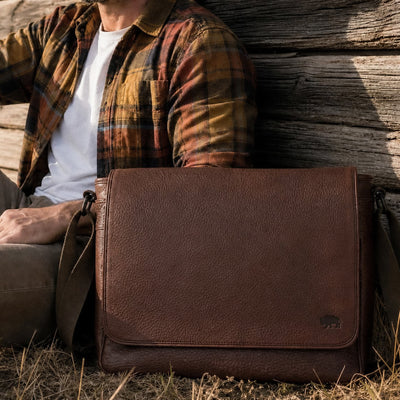 Person wearing a plaid shirt with a brown leather bag against a wooden wall.