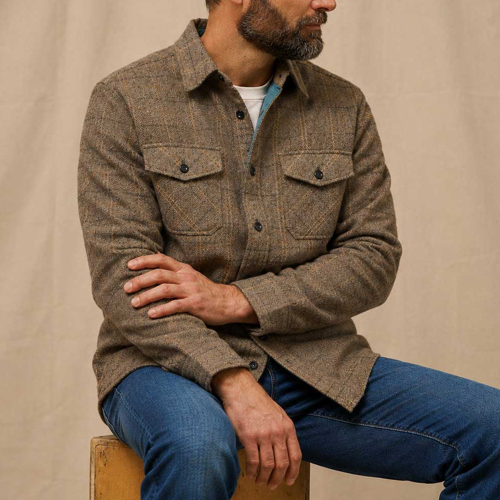 A man wearing a brown textured tweed button-up shirt and blue jeans, sitting on a wooden stool against a neutral beige fabric backdrop.