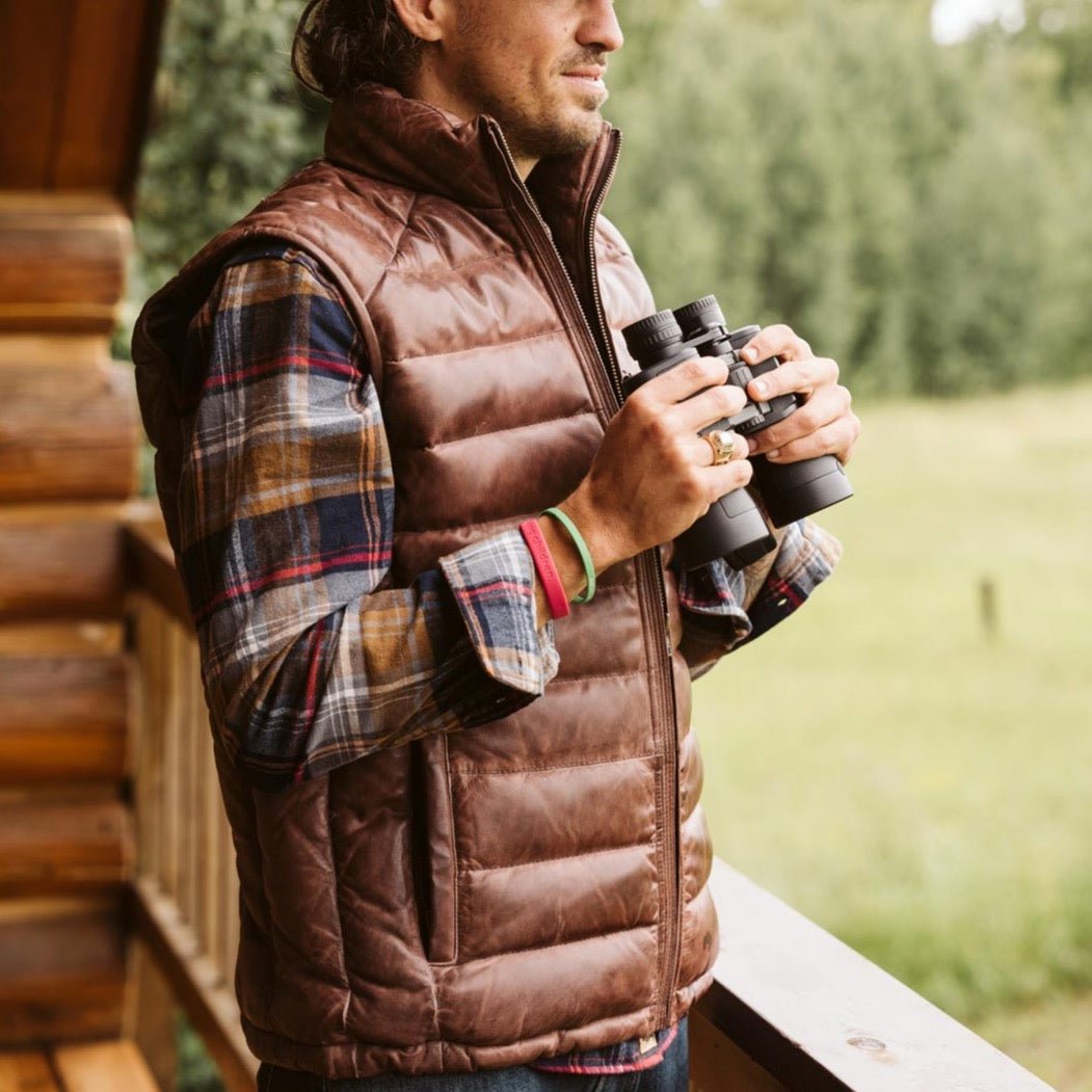 Man holding binoculars wearing the Bridger Leather Down Vest in Dark Brown over a plaid flannel