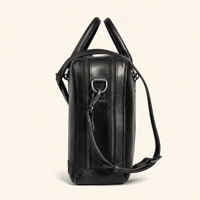 A black leather shoulder bag with dual top handles and a detachable shoulder strap, shown from a side profile view against a light cream background.