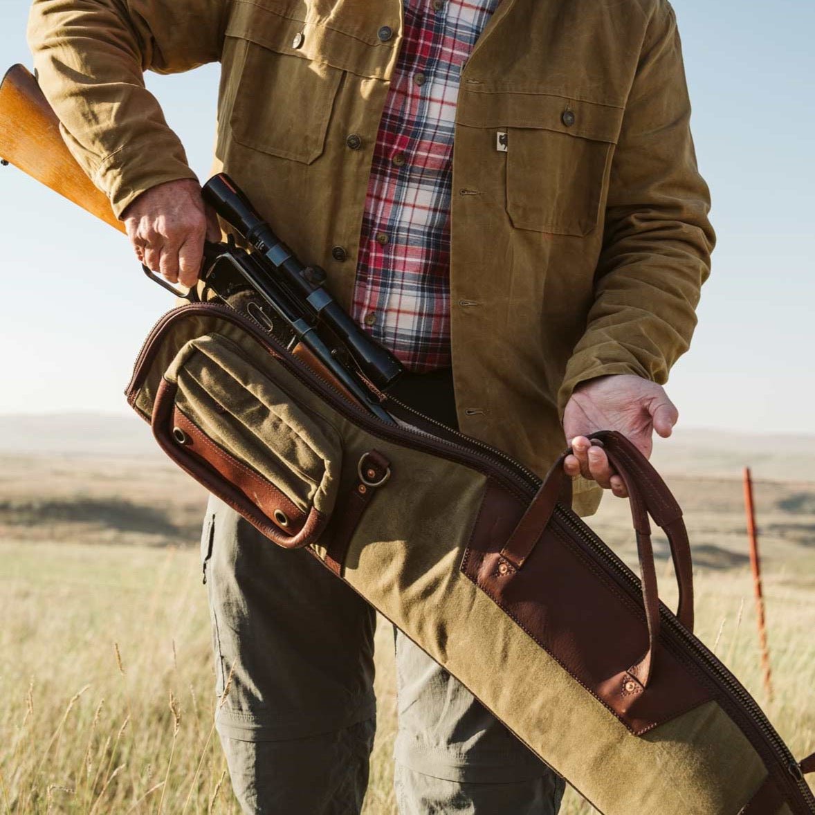 Hunter in a waxed cotton jacket carrying a Dakota waxed canvas and leather rifle case slung over one shoulder in an open field