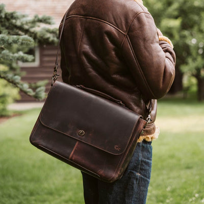 Man carrying Roosevelt Full-Grain Leather Messenger Bag in Dark Oak — back zipper pocket, worn crossbody in garden setting