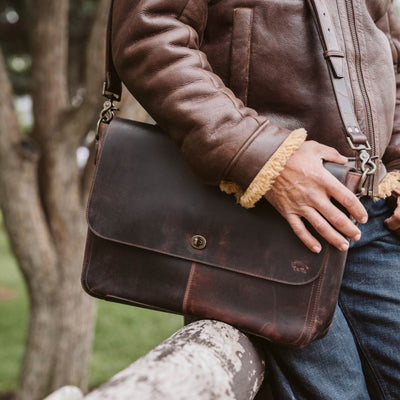 Man outdoors holding Roosevelt Full-Grain Leather Messenger Bag in Dark Oak — distressed leather, sturdy shoulder strap