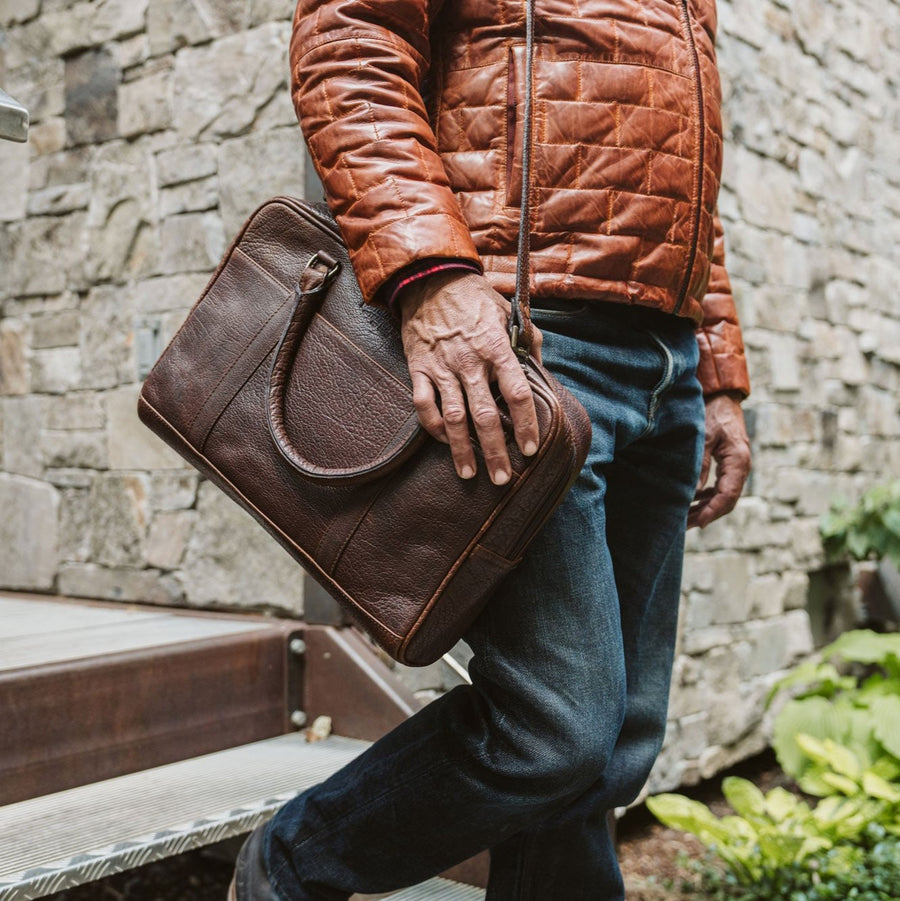 A brown leather laptop briefcase with a brass lock and a shoulder strap, set against a white background.