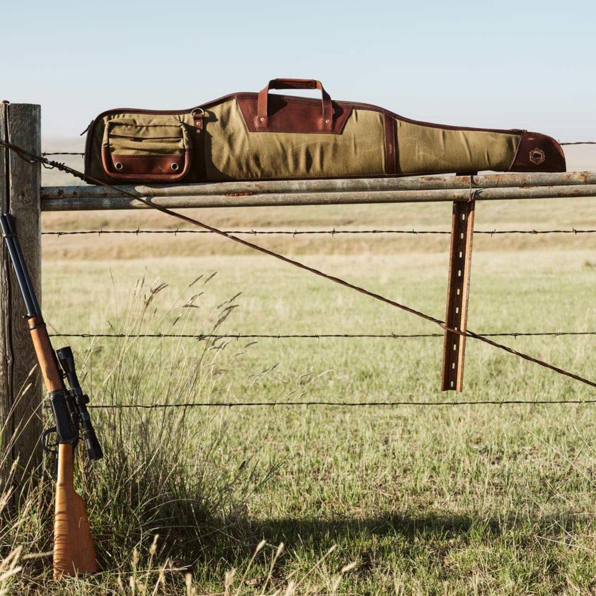 Waxed canvas and leather rifle case resting across a wire fence in a field of dry autumn grass