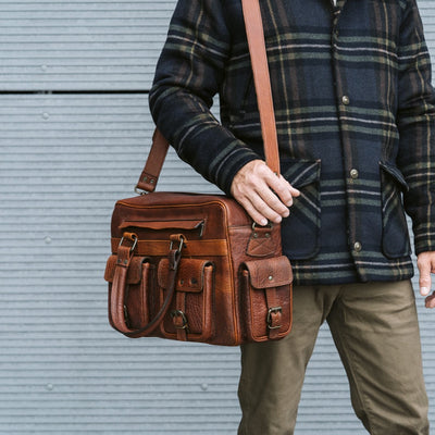 Man carrying vintage brown leather duffle bag with buckle straps and multiple pockets, wearing a plaid jacket, against a grey background.
