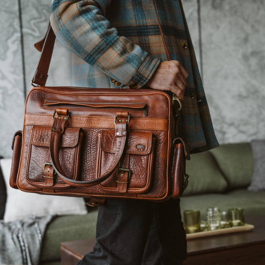 Young man holding a textured brown leather satchel with buckle closures, carrying it beside a chic, patterned jacket in a cozy indoor setting.