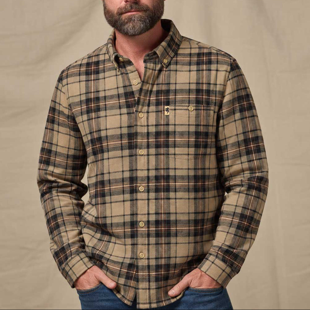 A man wearing a beige and brown plaid button-down shirt with blue jeans, standing against a neutral beige fabric backdrop.