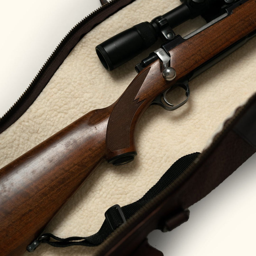 Scoped bolt action rifle with a walnut stock resting on the cream sherpa lining of an open leather rifle case
