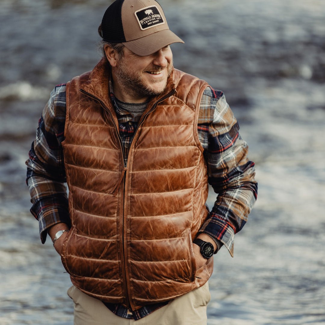 Man standing by a river wearing the Bridger Leather Down Vest in Light Brown over a plaid flannel