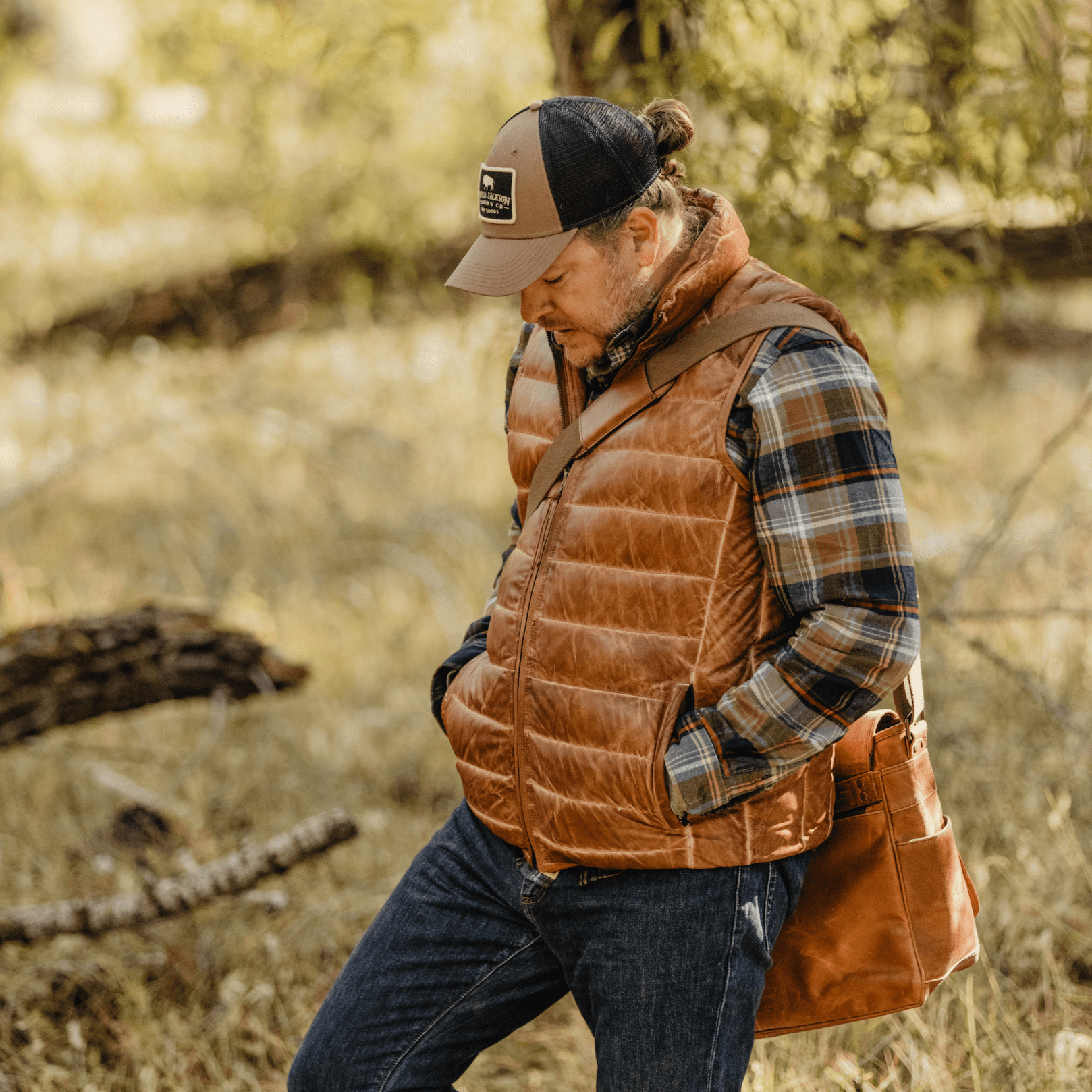 Man walking through a field wearing the Bridger Leather Down Vest in Light Brown over a flannel with a leather bag