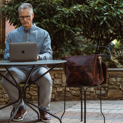 Dark oak leather messenger bag displayed next to a man working outdoors, showcasing its deep patina and robust build quality.