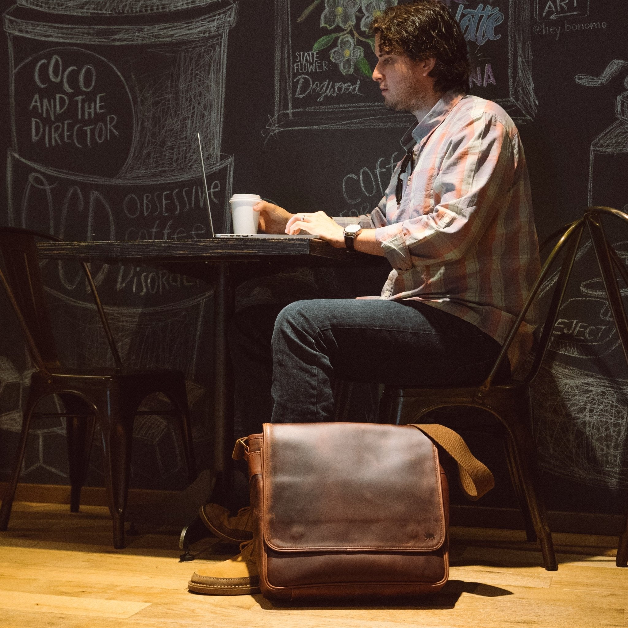 Roosevelt Buffalo Leather Satchel Messenger Bag in Dark Oak on a coffee shop floor beside a man working on a laptop