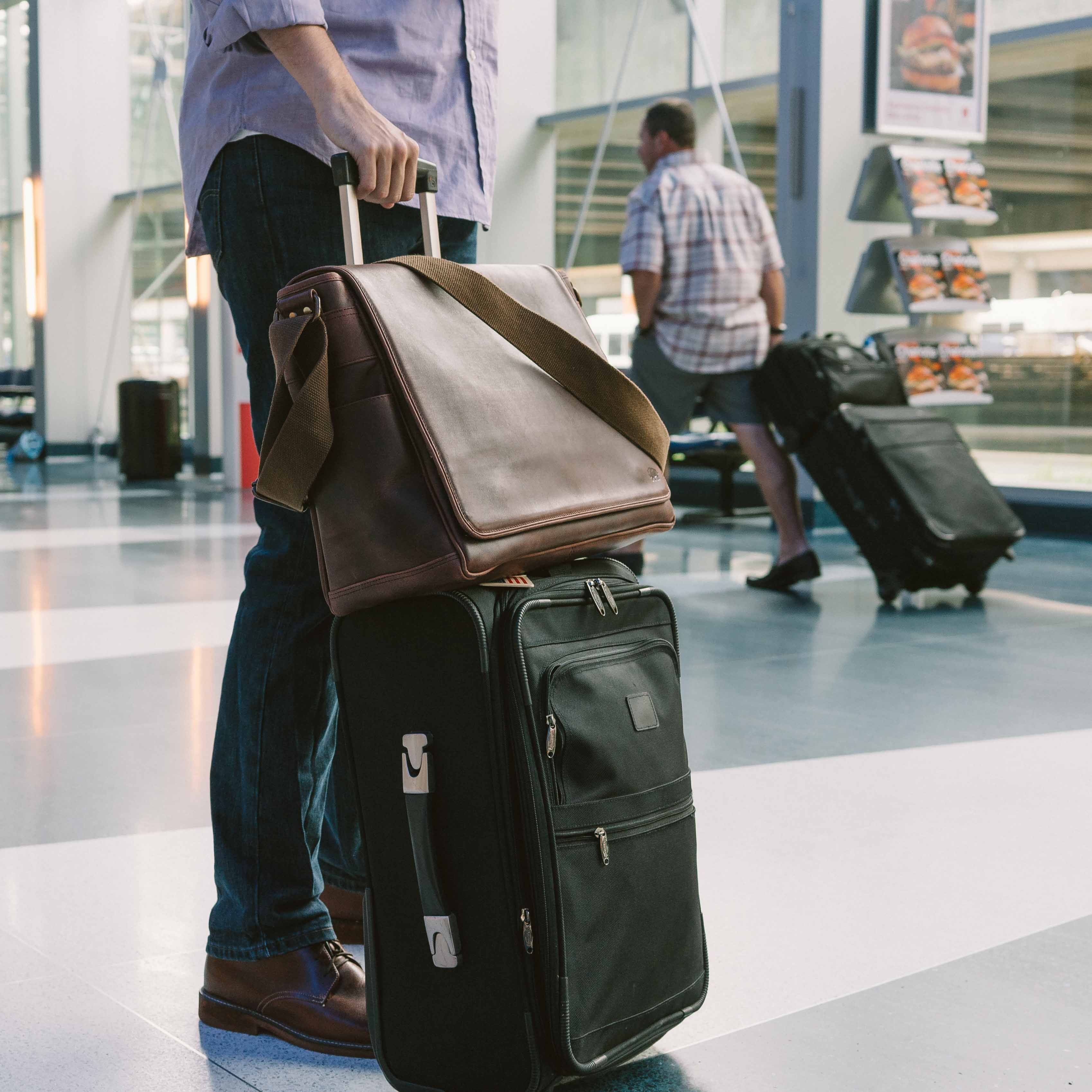 Roosevelt Satchel Messenger Bag Large in Dark Oak slid over a rolling suitcase handle in an airport