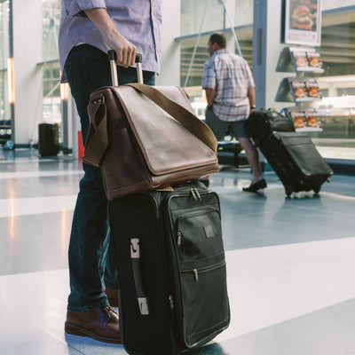 Stylish dark leather satchel with contrast strap, ideal for airport travel, easily attaches to rolling luggage handles.